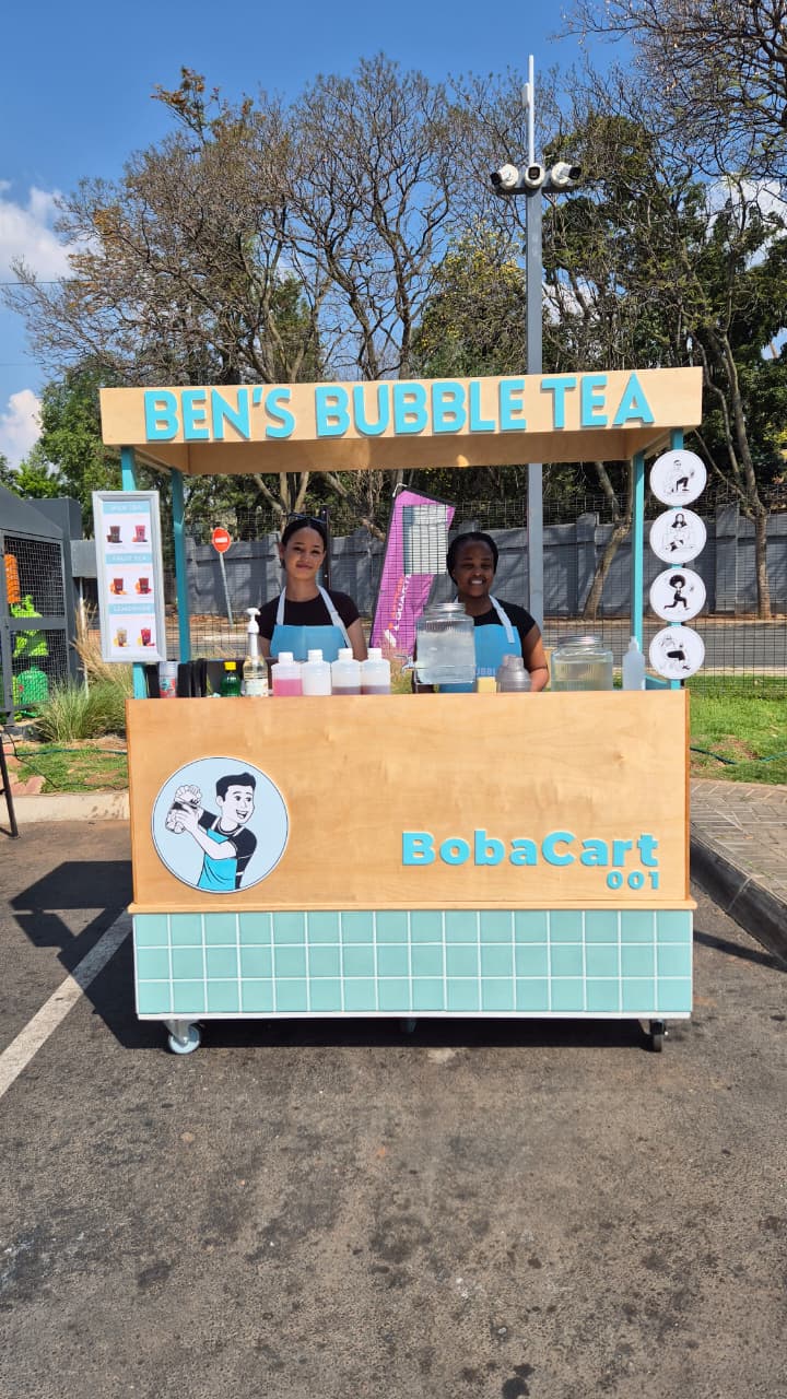 A mobile bubble tea cart with a light wood and teal tiled exterior, featuring the text 'BEN'S BUBBLE TEA' and 'BobaCart 001', with two people standing behind it outdoors on a paved area.