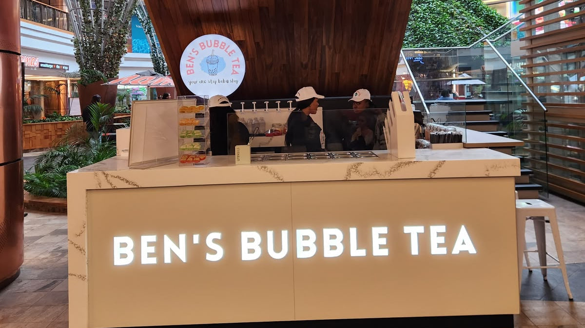 Ben's Bubble Tea storefront counter with illuminated white lettering, set in a modern indoor-outdoor space with wood paneling and greenery visible in the background.